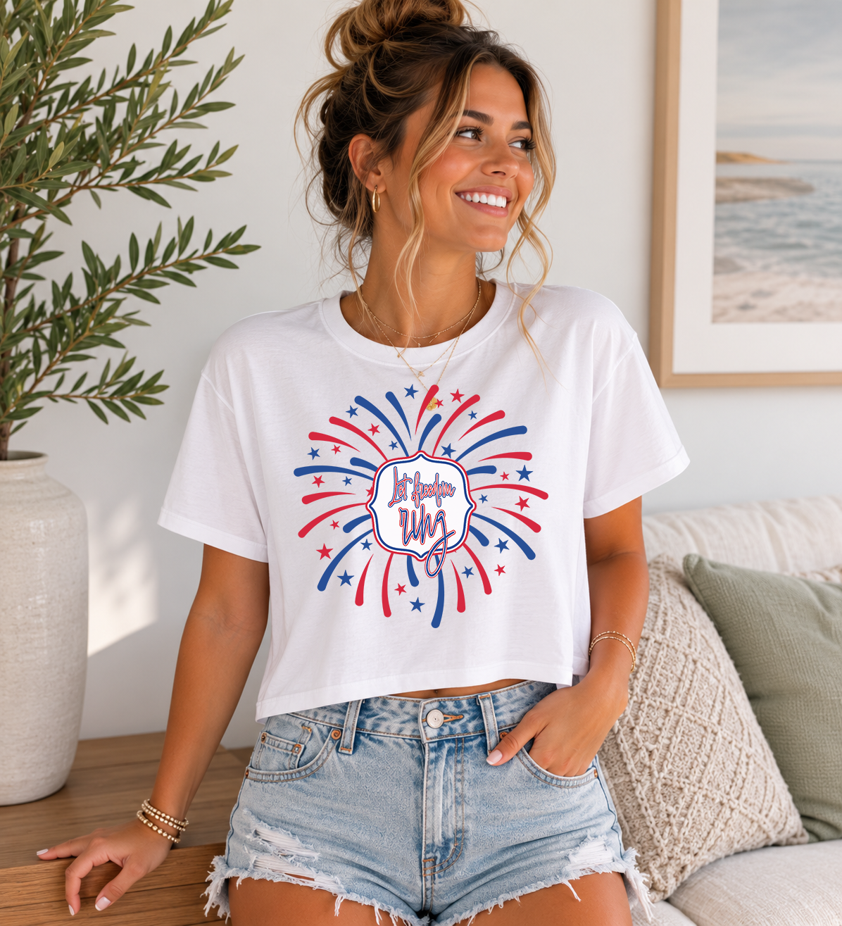 Adult female model wearing a white cropped graphic t-shirt featuring a red, white, and blue fireworks design surrounding the phrase “Let Freedom Ring,” styled with distressed denim shorts in a bright neutral indoor setting.