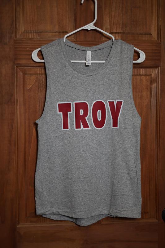 Heather gray muscle tank featuring bold red and white TROY lettering on the front, displayed on a hanger against a wooden door background.