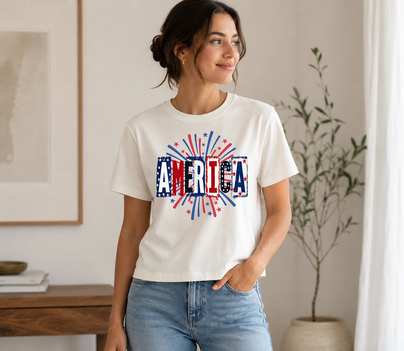 Adult female model wearing a white graphic t-shirt featuring a distressed America design with red, white, and blue fireworks accents. Styled with light wash jeans in a neutral indoor setting.