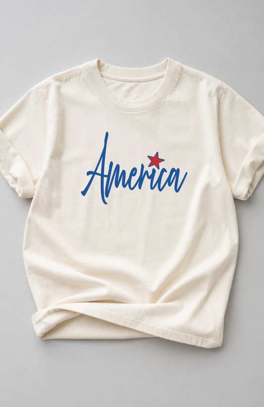 Ivory t-shirt with blue script “America” text and a small red star detail, styled in a minimal flat lay on a neutral background