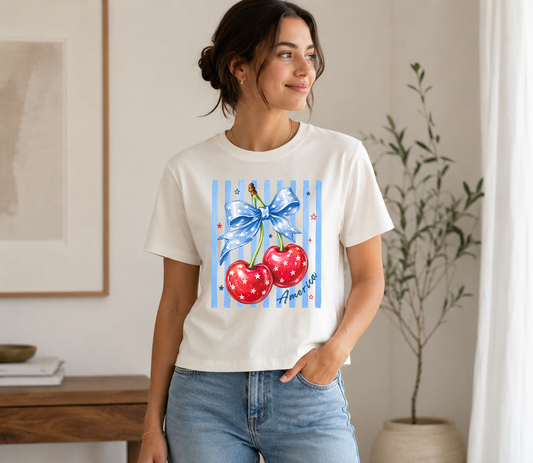 Adult female model wearing an ivory graphic t-shirt featuring patriotic red cherries tied with a blue star-print bow, accented with blue stripes and Americana-inspired details, paired with light wash jeans in a neutral indoor setting.