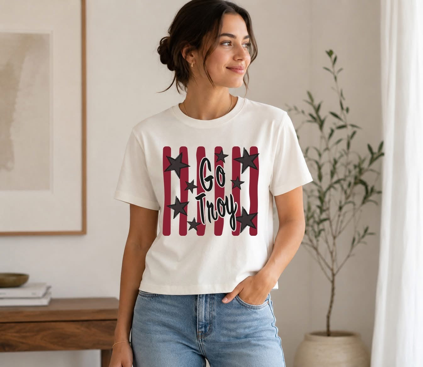Lifestyle photo of a woman wearing a white short-sleeve t-shirt with cardinal red vertical stripes, black stars, and black script lettering that reads Go Troy.