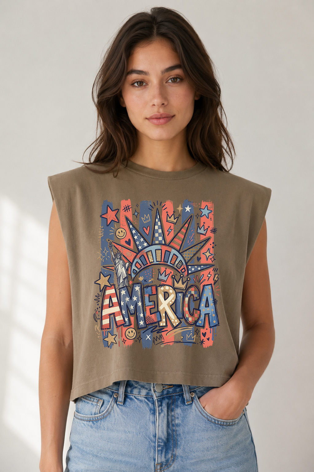 Adult female model wearing a cropped taupe muscle tank featuring a colorful doodle-style America graphic with patriotic stars, crowns, Statue of Liberty accents, and bold lettering, paired with light wash jeans in a neutral setting.
