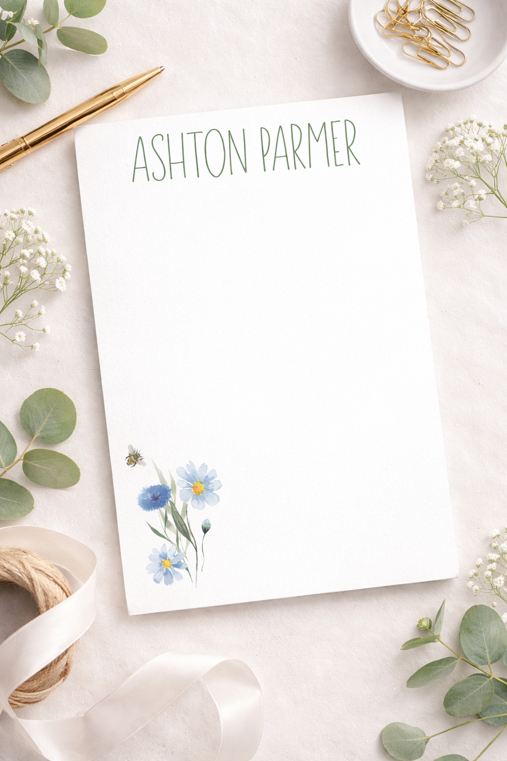 Notepad with floral design and 'Ashton Parmer' (customizable) text on a decorative surface with flowers and ribbons.