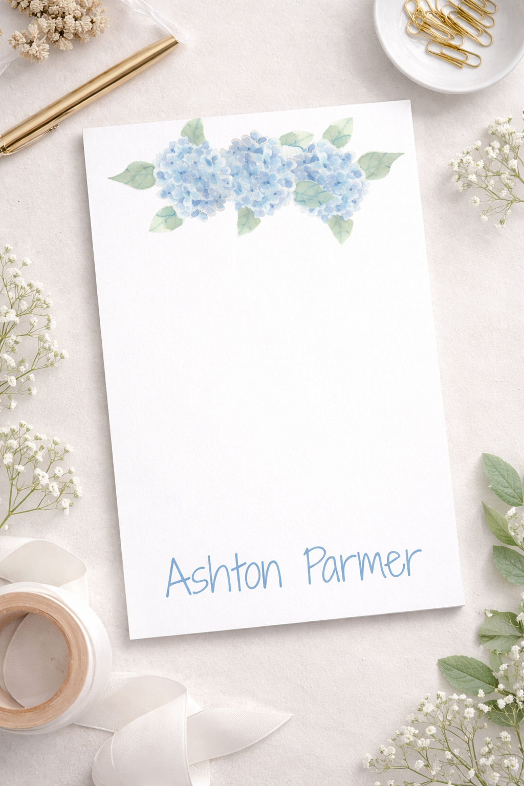 White notepad with blue hydrangea design at top and 'customizable name' at bottom surrounded by decorative elements on a light surface for background.