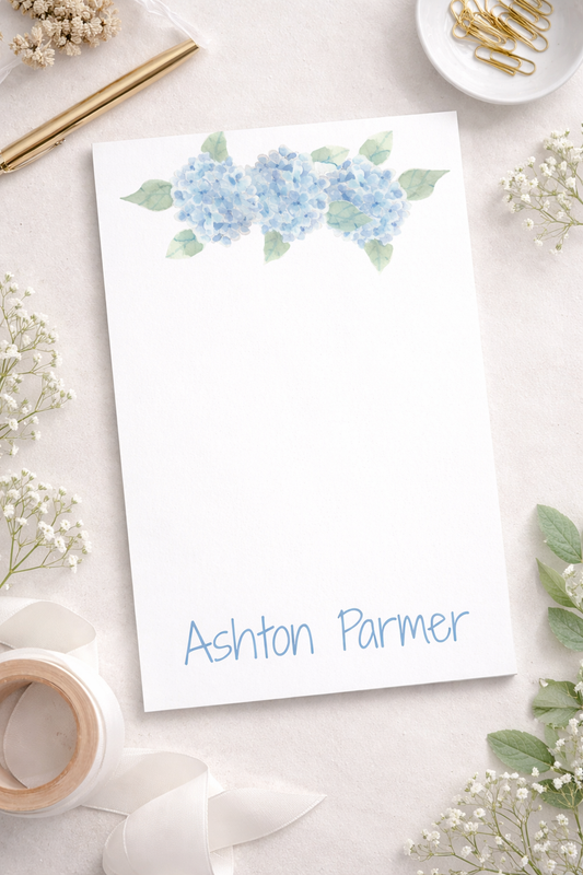 White notepad with blue hydrangea design at top and 'customizable name' at bottom surrounded by decorative elements on a light surface for background.