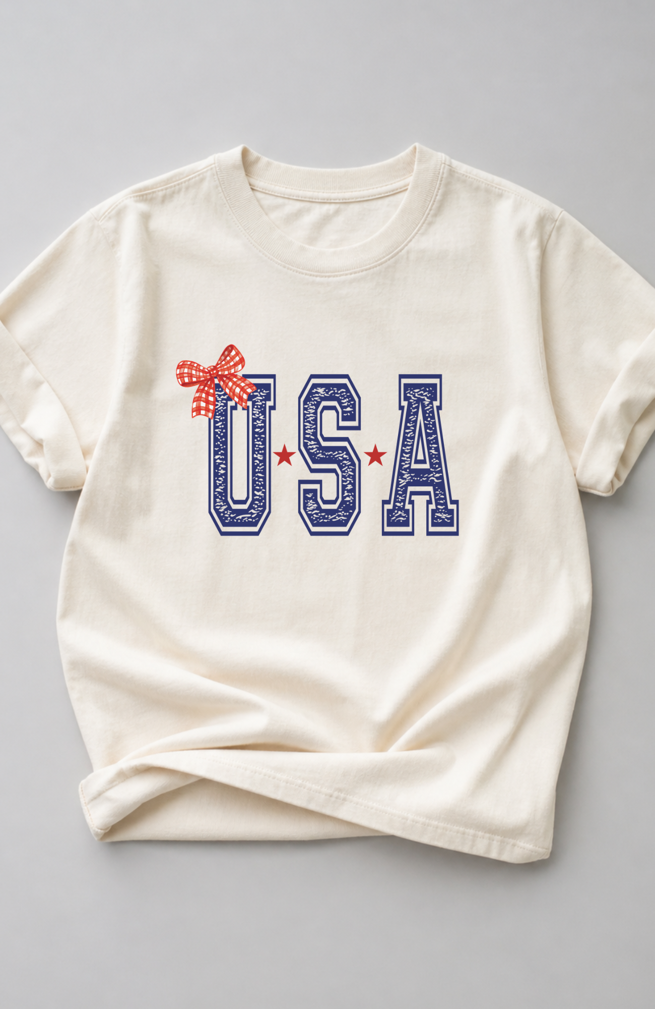 Ivory t-shirt with bold blue “USA” lettering, red star accents, and a red gingham bow detail, styled in a minimal flat lay on a neutral background