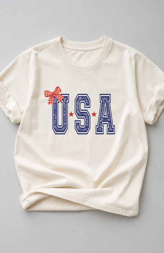 Ivory t-shirt with bold blue “USA” lettering, red star accents, and a red gingham bow detail, styled in a minimal flat lay on a neutral background