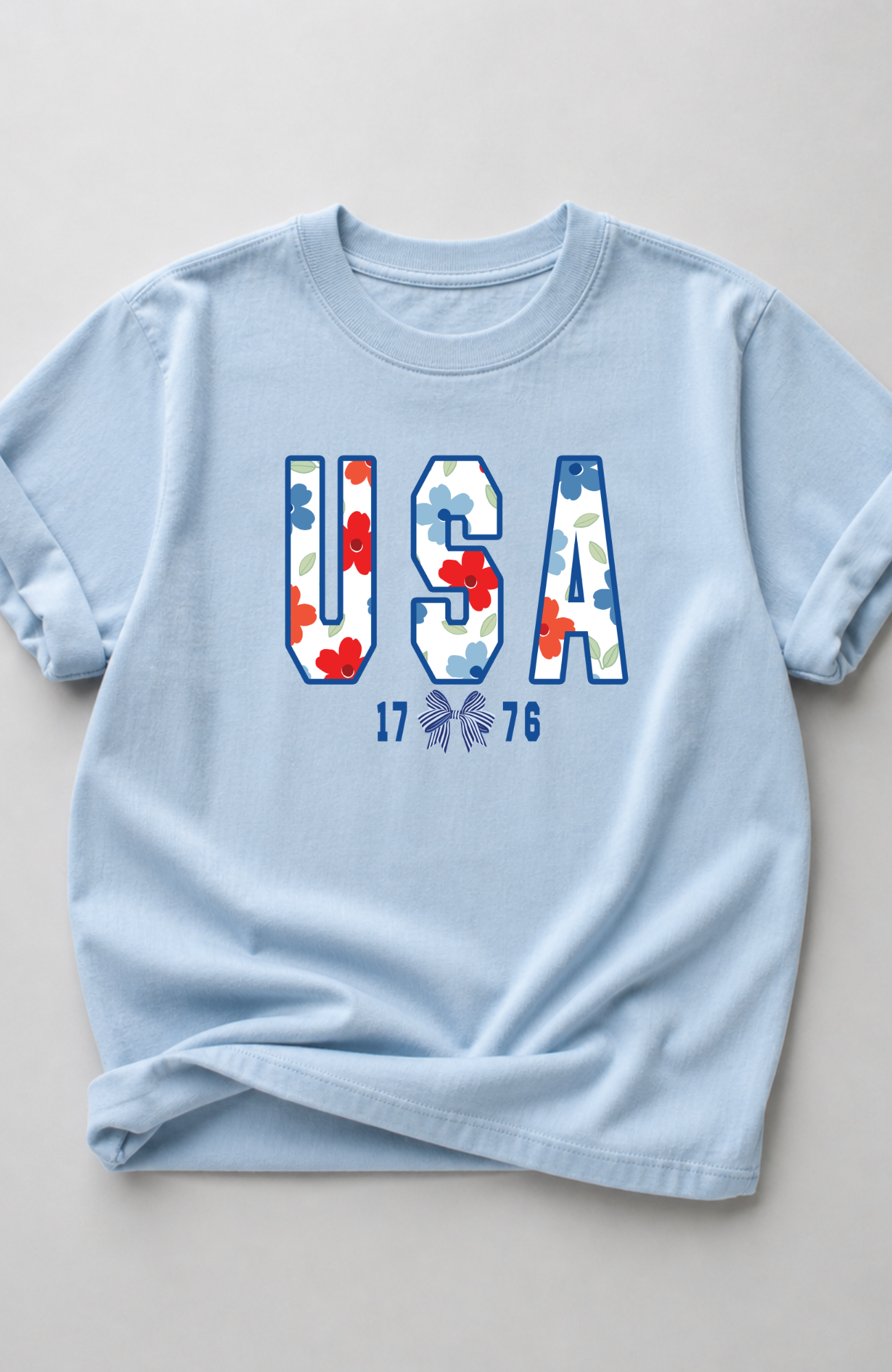 Light blue t-shirt with large “USA” lettering filled with red, white, and blue floral pattern and small “1776” detail, styled in a minimal flat lay on a neutral background