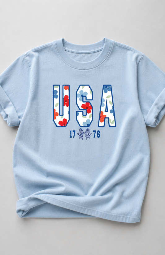 Light blue t-shirt with large “USA” lettering filled with red, white, and blue floral pattern and small “1776” detail, styled in a minimal flat lay on a neutral background