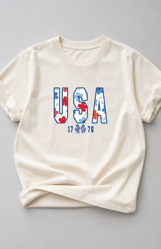 Ivory t-shirt with large “USA” lettering filled with red, white, and blue floral pattern and small “1776” detail, styled in a minimal flat lay on a neutral background