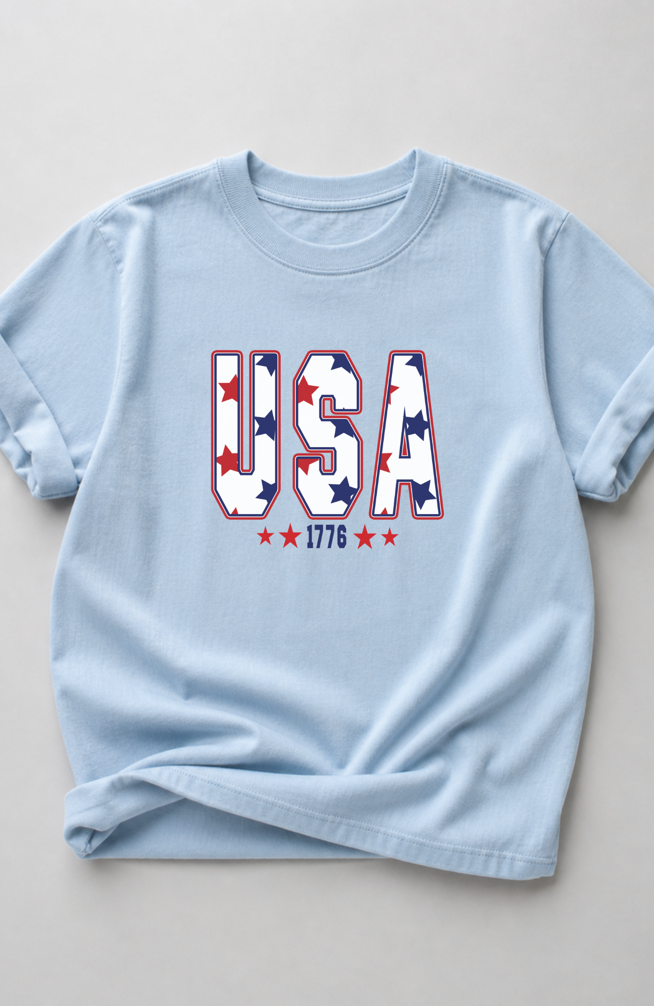 Light blue t-shirt with bold “USA” lettering filled with red and blue stars and small “1776” detail, styled in a minimal flat lay on a neutral background
