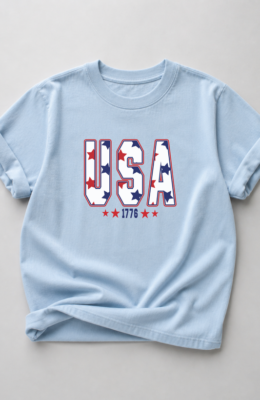 Light blue t-shirt with bold “USA” lettering filled with red and blue stars and small “1776” detail, styled in a minimal flat lay on a neutral background