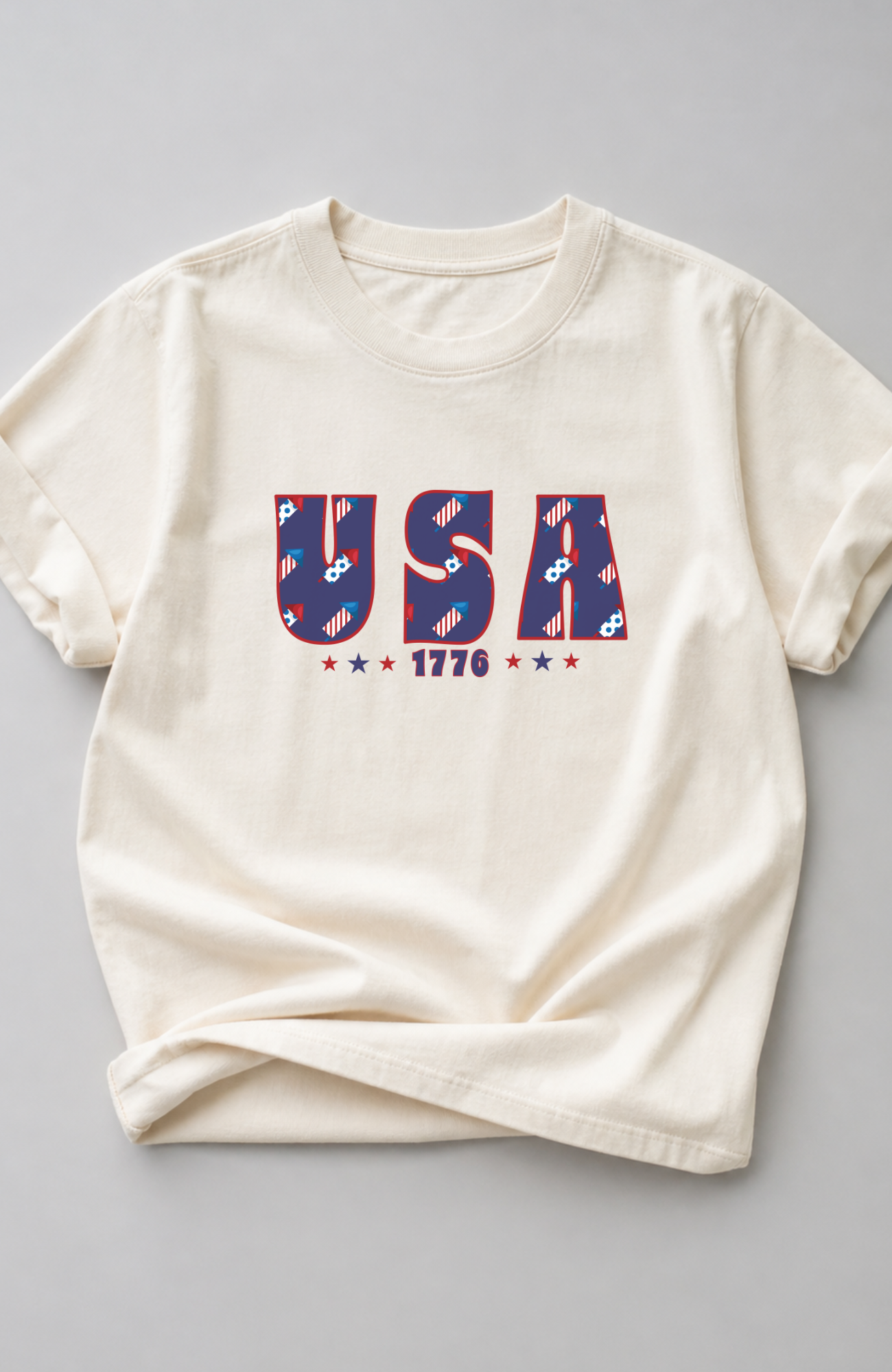 Ivory t-shirt with bold “USA 1776” graphic in red, white, and blue patterned lettering, styled in a minimal flat lay on a neutral background